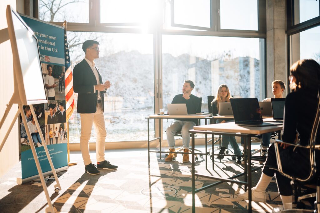 US-Studienberater Jonas von Epro 360 präsentiert vor einer Gruppe Studierender in einem modernen Seminarraum mit US-Flagge und Präsentationsmaterialien.