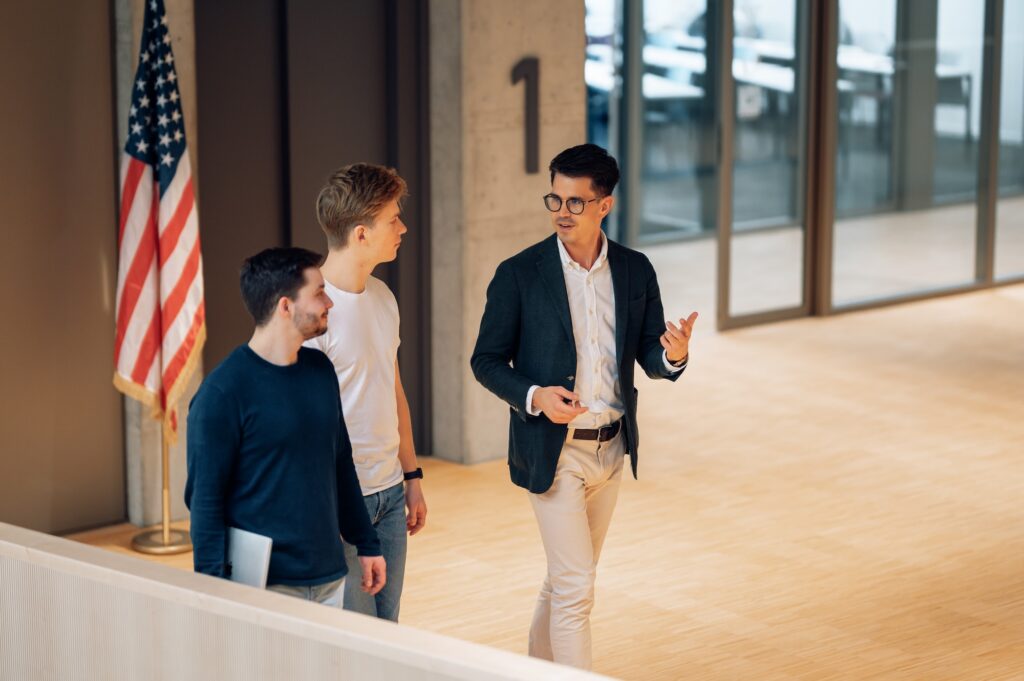 Dozent spricht mit zwei Studierenden in moderner Universitätshalle neben einer US-Flagge.